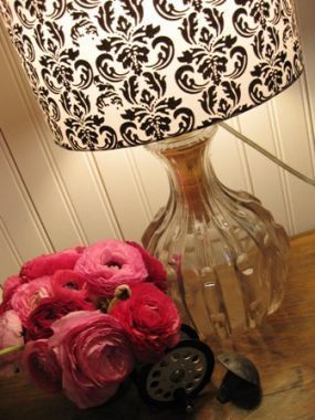 A vintage decanter has been turned into a lamp with a printed shade and is displayed next to an arrangement of pink flowers.