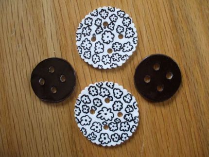 Four buttons, two small black ones, two large white ones with black flowers on them sit on a wood table.