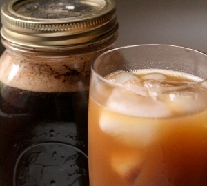 Some iced coffee in a clear glass with some ice sits next to a ball jar of coffee.
