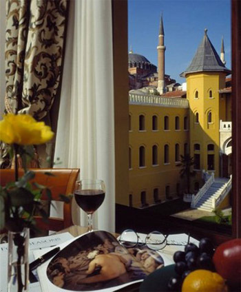 Food, flowers and wine on a table next to curtains and a window facing a yellowish chateau.