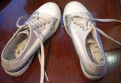 A pair of shabby white low cut sneakers, with laces undone, sitting on a wood floor.