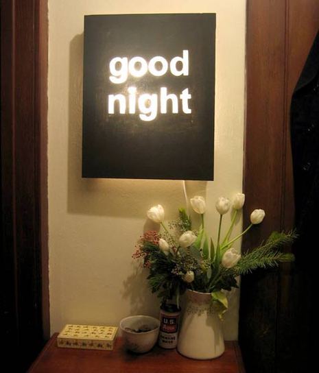 A glowing sign with a black background and white letters that says "goodnight" hangs on a white wall, with a vase of white flowers underneath.