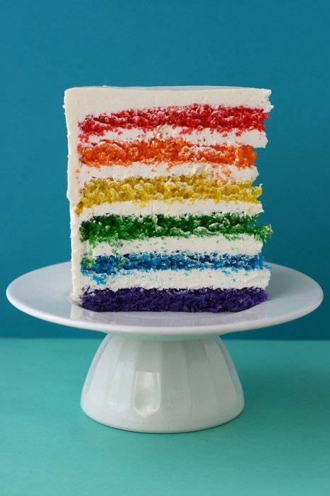 A large slice of white frosted cake with layers resembling a rainbow is on a cake plate.