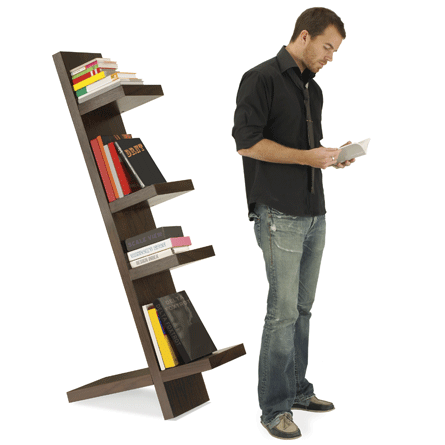 A man stands reading a book in front of a tilted backwards bookshelf.