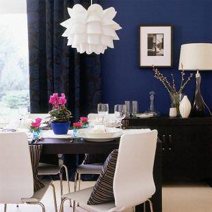 A white petal-like lamp sits above a dining table that is set for dinner in a royal blue painted dining room.
