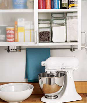 A white kitchenmaid mixer with a silver bowl next to white mixing bowls in front of a blue towel and under a shelf with flour, sugar and staples in it.