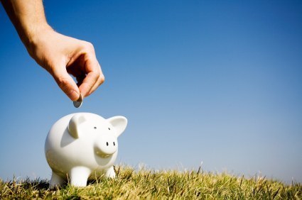 A hand is placing a coin in a white piggy bank in the grass.