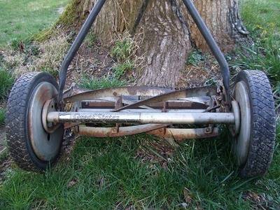 An old 5 blade rotary wheel lawn mower sitting against a tree.