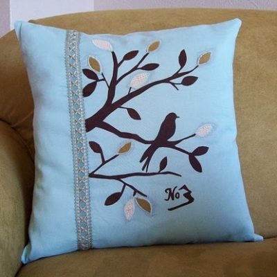 A light blue decorative pillow that has brown leaves and branches, with the silhouette of a bird, sitting against a tan couch.