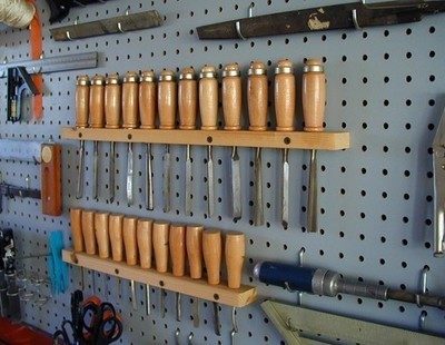Various wood handled chisels are hung on a wall with other tools nearby