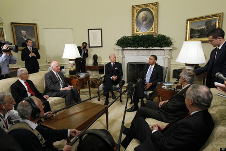 Barack Obama and Joe Biden in a room in the Oval Office meeting with a bunch of other people.