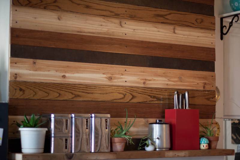 A shelf below planks of different colored woods containg three silver containers three plants and a red knife holder.