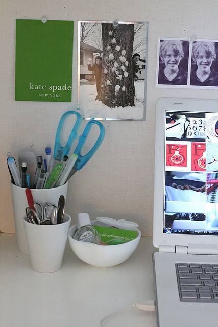 Part of a laptop, a couple of white office supplies holders filled with pens, scissors, tape and a coule of pictures on the wall.