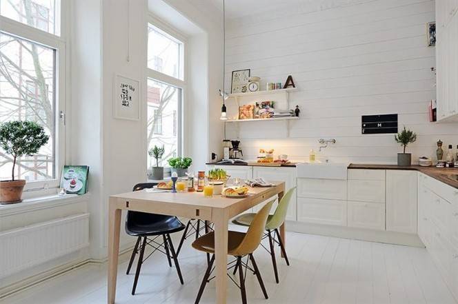 A wood dining table with various colored dining chairs sits in a white kitchen