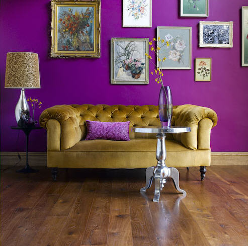 A mustard yellow classic sofa sits in front of a purple wall with picture frames decorating it