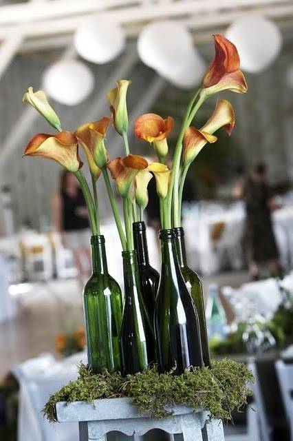 Orange and yellow flowers are arranged in green wine bottles on a table covered in Moss