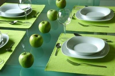 Dining table decorated with green apples, glasses, green table mats with plates, bowls, spoons and forks at top.