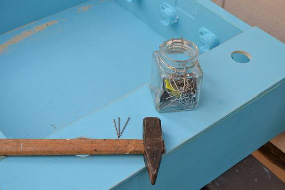 A mason jar full of nails and other assorted trinkets next to a hammer on a blue shelf.