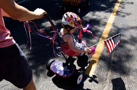 An adult pushing a young boy in a stroller who is holding a miniature American flag.