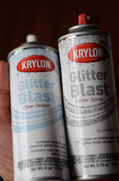 Glitter spray and glitter sealer spray cans are held in someone's hand.