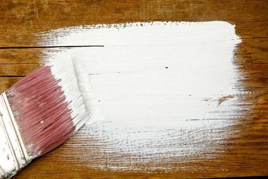 A paintbrush with white paint brushing wood.