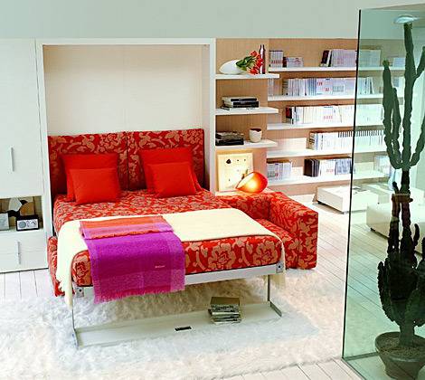 A pull down bed made in a red and gold cover with red pillows and a tan throw with a tall cactus behind green glass.