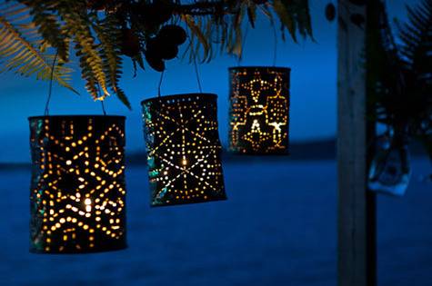 Three lamps, black cylinders delightfully punctured to make starry designs, hang from a tree overlooking the ocean at dawn.