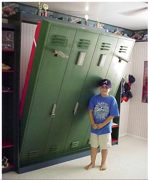 A young boy in a blue shirt and dark ball cap is standing next to green lockers that double as a pull down bed.