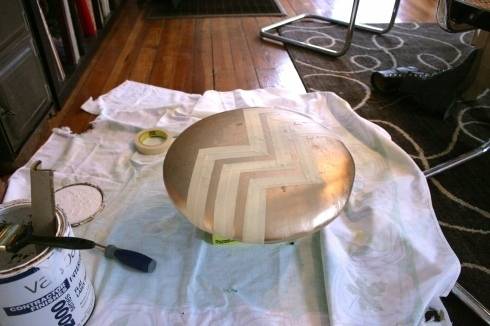 A round metallic cushion has masking tape over it in a geometric design and is next to a can of paint and paintbrush.