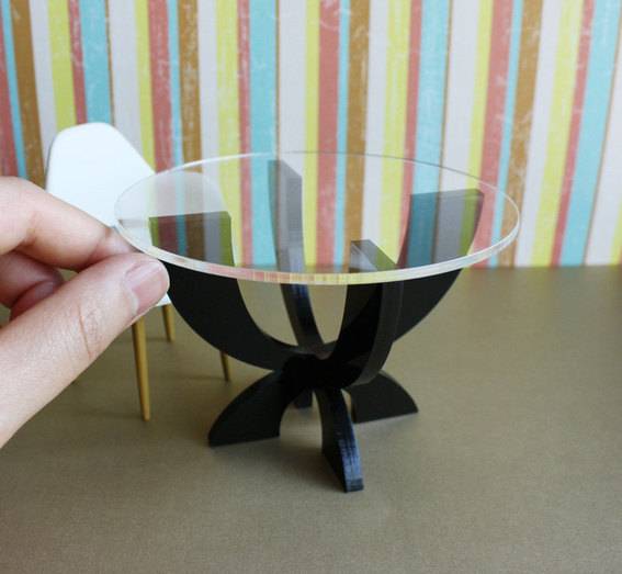 A round miniature glass top table, being placed by hand, in front of a miniature chair.