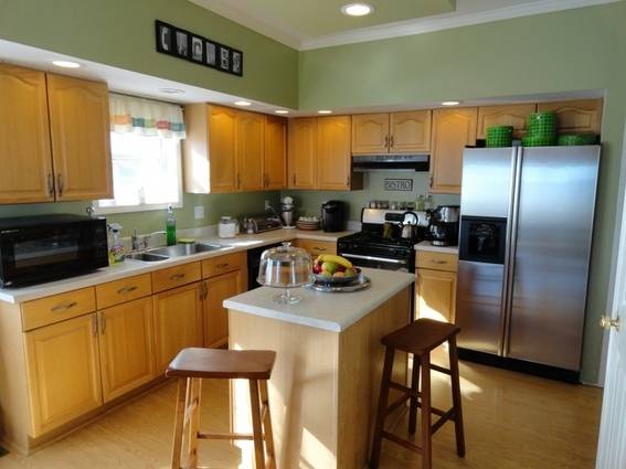 Neat kitchen with honey-colored wood cabinets, two stools around a center island, and a large stainless steel refrigerator.