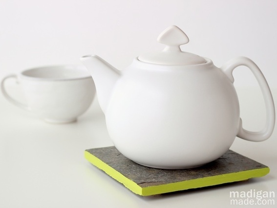 A white ceramic tea pot with lid sits on a yellow and grey  square coaster to the right of a matching ceramic tea cup.