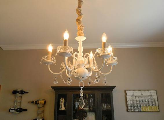 A six arm white chandelier with crystals hanging down and lit candle shaped lights in a tan room.