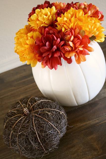 A white vase with yellow and maroon flowers next to a black mini pumpkin.