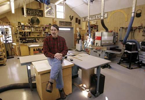 A man sits cross-legged on a counter in a full, but neat garage workshop.