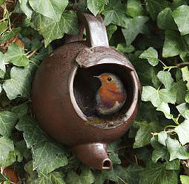 A bird resting in a brown tea kettle surrounded by green leaves.