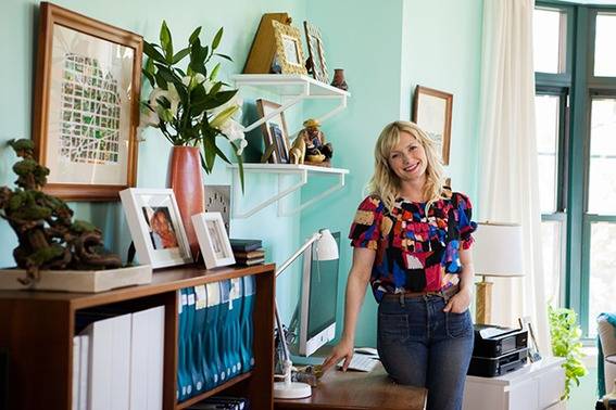 A woman stands in an office that has a desk, electronics, binders, and photos.