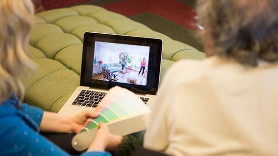 Two women look at a laptop screen while one of them holds a palette of paint colors.