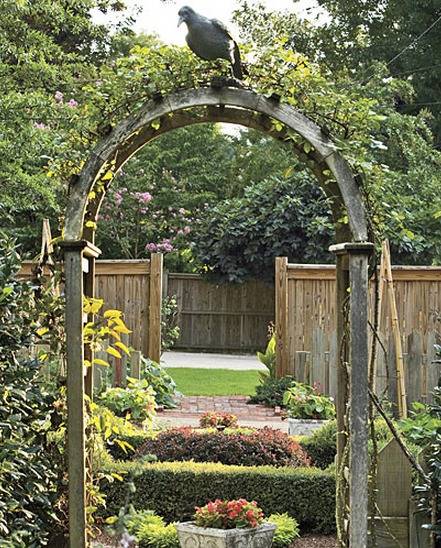 A wooden garden arch is at one side of a yard and a privacy fence is at the other side.