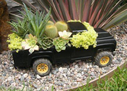 A miniature decorative pickup truck has been filled with plants and is sitting next to a larger plant in a garden.