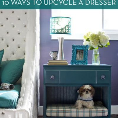 An old dresser has had some drawers removed and a small dog bed added for a fun dog hideout that doubles as a nightstand with a lamp, vase, and framed photo on top.