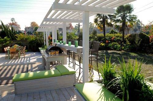 A pergola covers a bar next to a dining table with chairs and other seating on a patio.