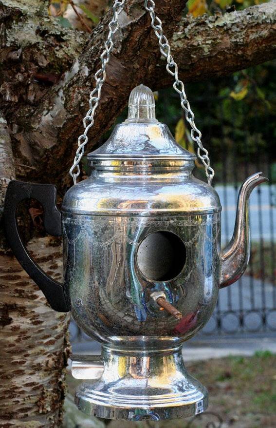 A silver teapot is hanging from a chain in a tree.