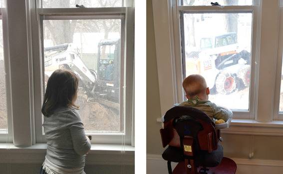 Girl looking out window at a small tractor next to boy sitting in chair looking out window.
