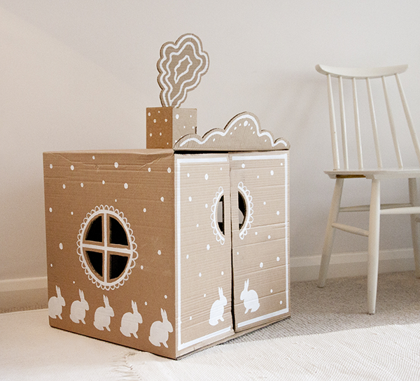 A pink cardboard playhouse, decorated with white bunnies, nestles beside a chair.