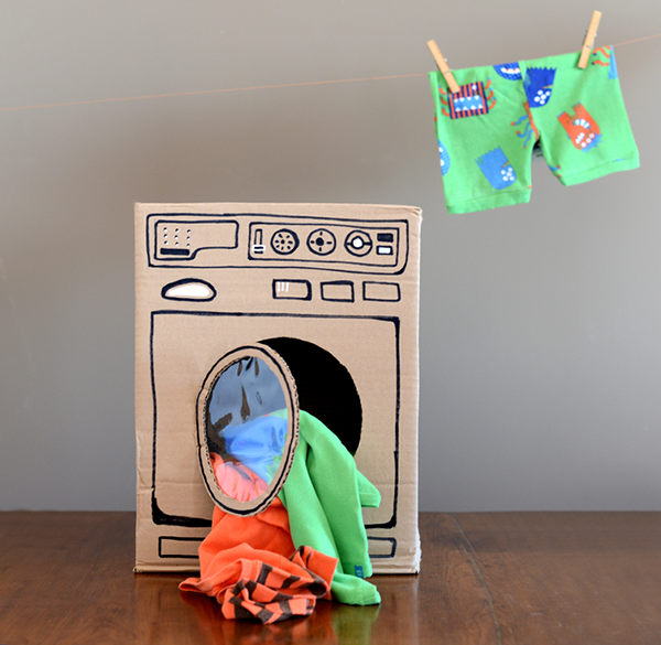 Colorful clothing is hanging out of a cardboard washing machine.