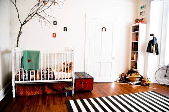 A white crib sits under a tree in a room with a wooden floor and white and black striped rug.
