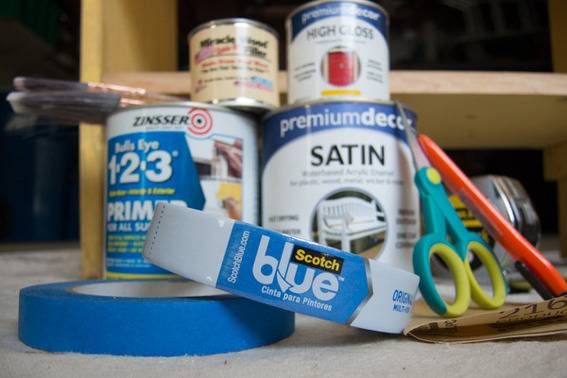 A few cans of paint, a couple of paint brushes, some marking tape, a measuring ruler, a pair of scissors and a pen sit in front of a wooden cabinet.