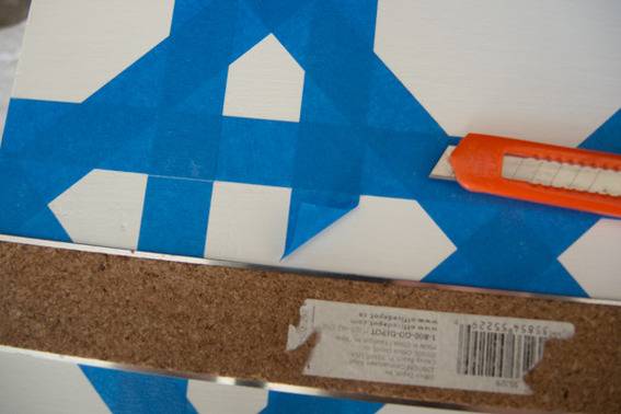 An orange boxcutter sits next to a ruler on white paper with blue painting tape on it.
