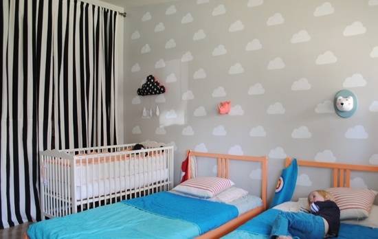A white crib and two beds with blue sheets are in a room with black and white curtains.